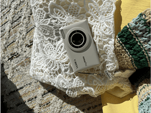 Charger l&#39;image dans la galerie, Un appareil photo sur une surface de dentelle blanche, avec un objet jaune et une couverture à motifs.
